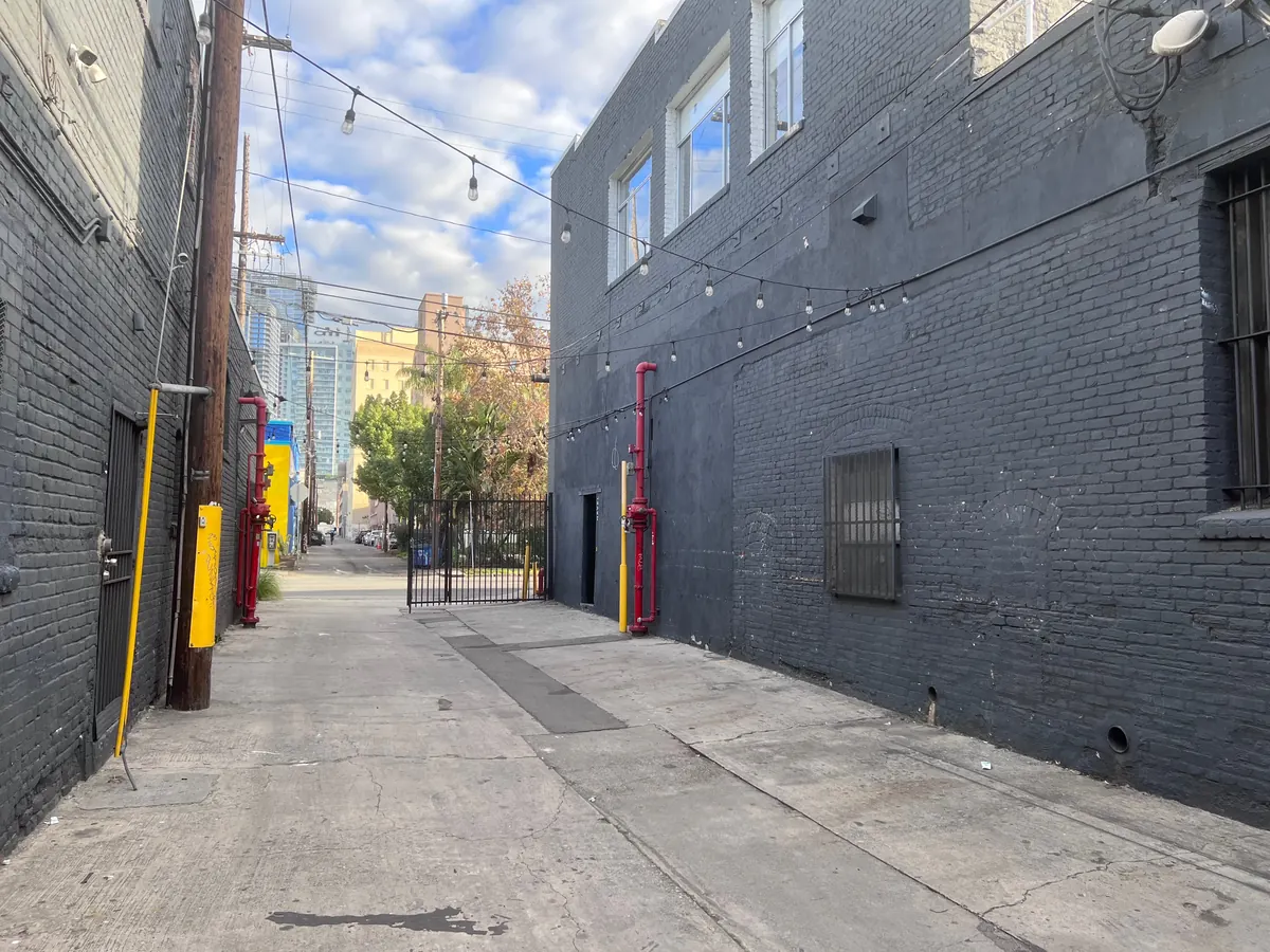 The empty alley in daylight, with string lights overhead and downtown towers beyond the gate