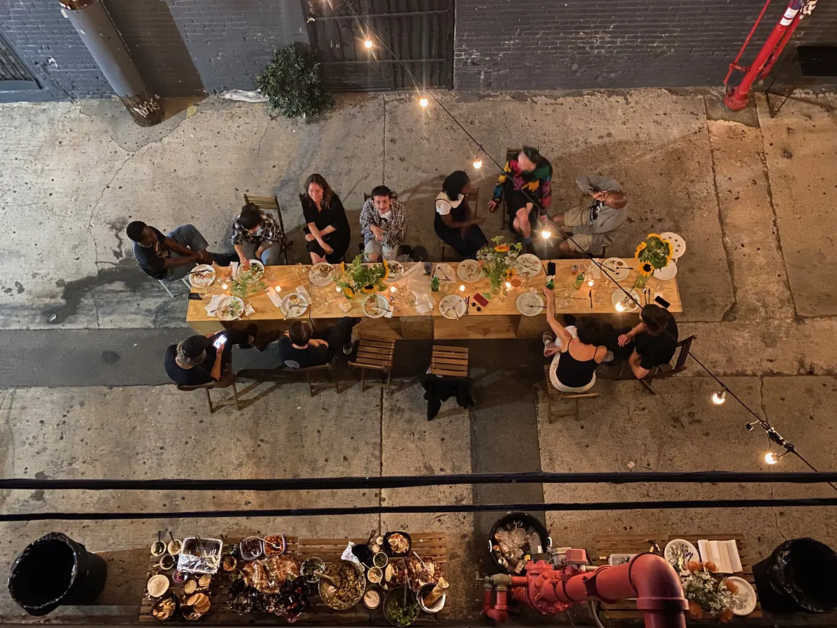 A long table of dinner guests in the alley, seen from above, under string lights